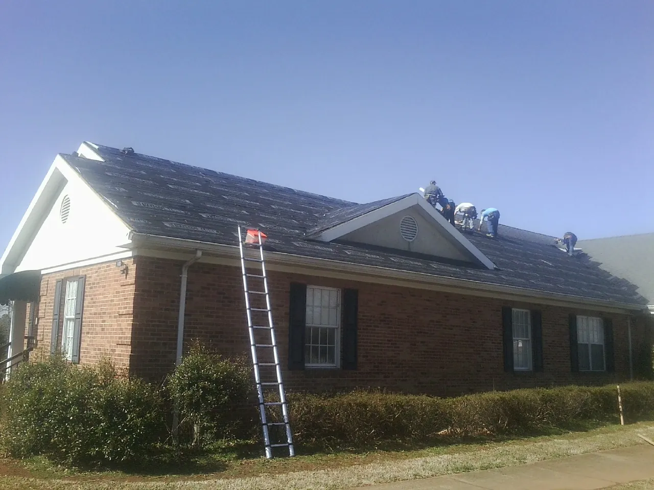 Crew tearing shingles off a brick ranch roof with a ladder set — Emergency Roof Repair work by our crew in Roosevelt
